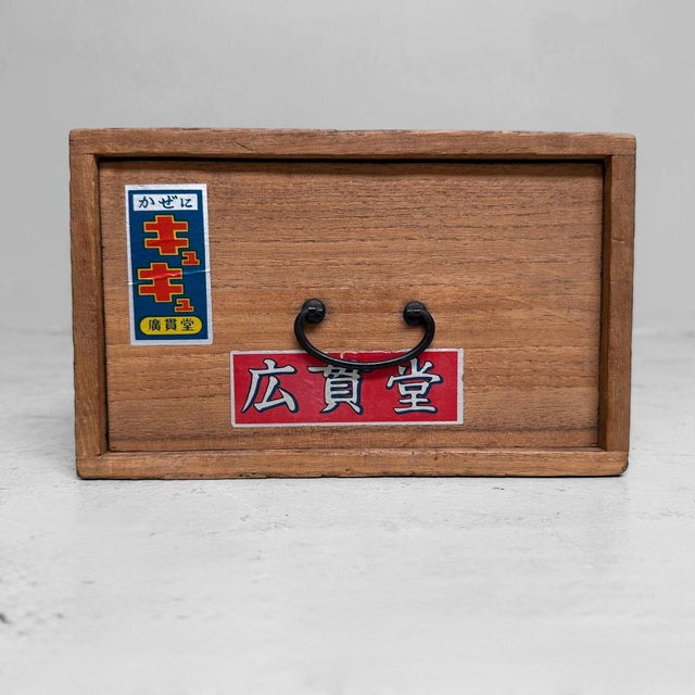 A small Japanese wooden pharmacy cabinet with one drawer, a metal pull handle, and original medicine advertising labels on...