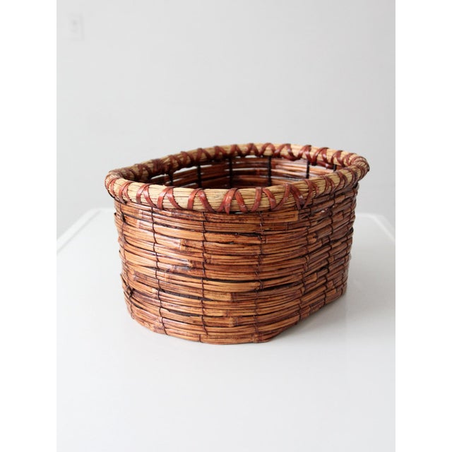 A vintage woven basket with an oval form and a warm honey-brown tone, finished with a deep red wrapped rim that adds...