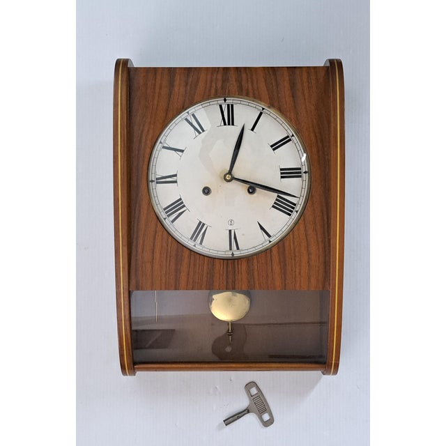 A beautifully preserved vintage mechanical pendulum wall clock housed in a warm teak wood case with softly curved side...
