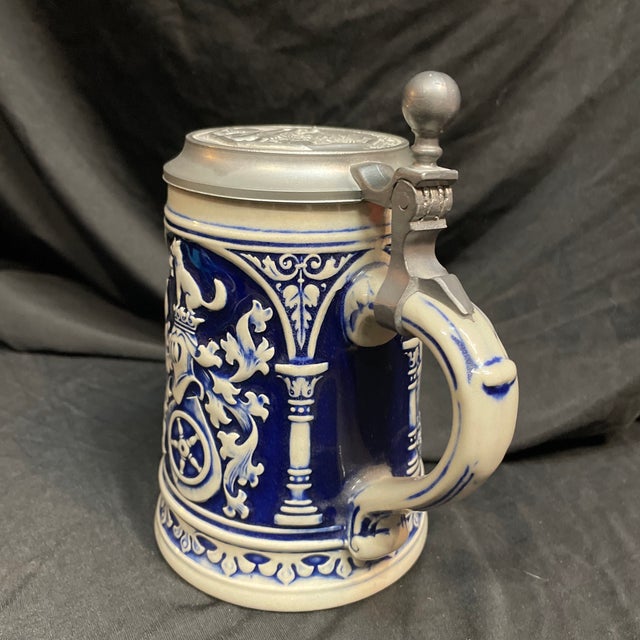 A ceramic German drinking vessel with ornamented pewter lid, decorated with Bavarian castles, an antique militia, and a...