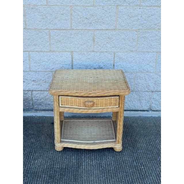 Vintage wicker nightstand in a natural beige-tan finish, featuring a woven rectangular top, single drawer with a matching...