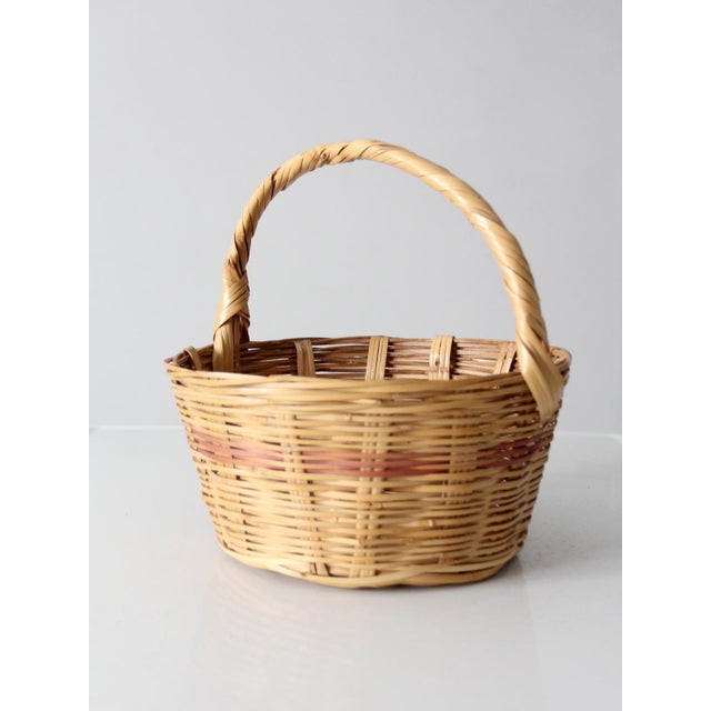 This vintage woven gathering basket pairs warm natural cane with a subtle rosy pink band around the body. Its rounded...