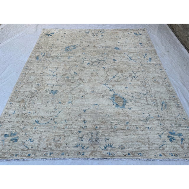 An ivory field with soft blue floral vines defines this handmade Oushak wool rug. Taupe and sand-colored leaves weave...