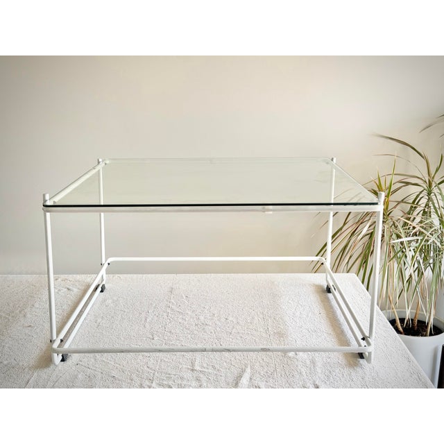 A clean and architectural tubular steel coffee table featuring a white enamel-finished frame and floating glass top. The...