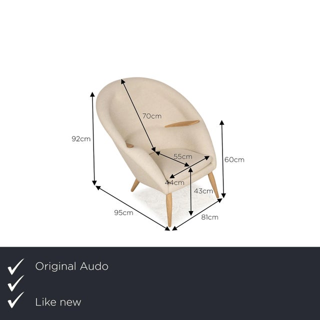 The Oda armchair from Audo is covered in a beige fabric and stands on brown wooden feet. It is characterized by its...