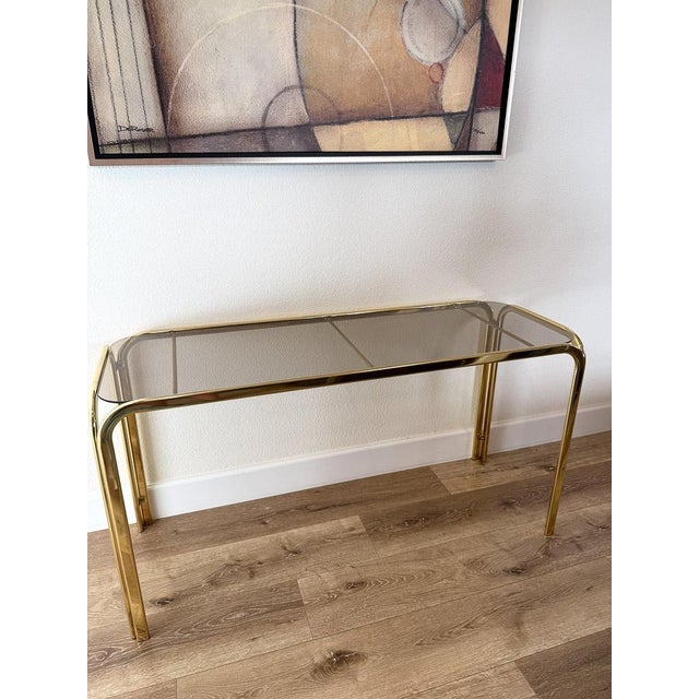 Elegant vintage Hollywood Regency console table featuring a polished brass frame with waterfall edges and inset smoked...