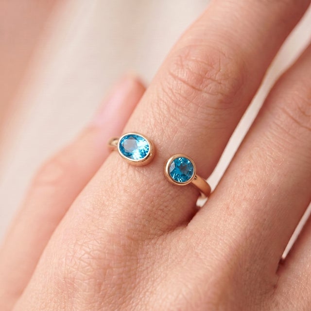 A bold, contemporary open-cuff ring featuring two brilliant blue topaz stones — an oval-cut stone paired with a round-cut...