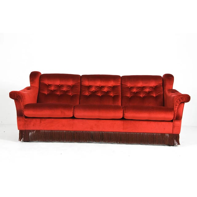 Three-seat sofa upholstered in red velour with corded trim, raised on beechwood feet. The sofa has a low, wide profile...