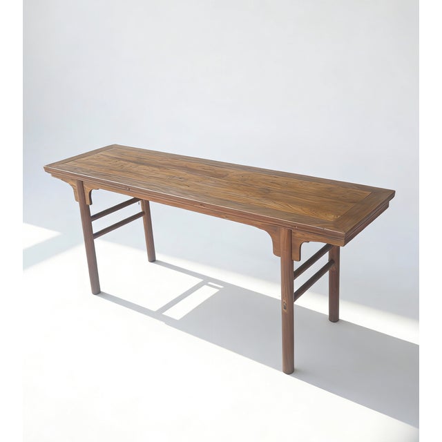 This long, slender console table is crafted from solid elm wood, featuring a rectangular top with a visible natural grain....