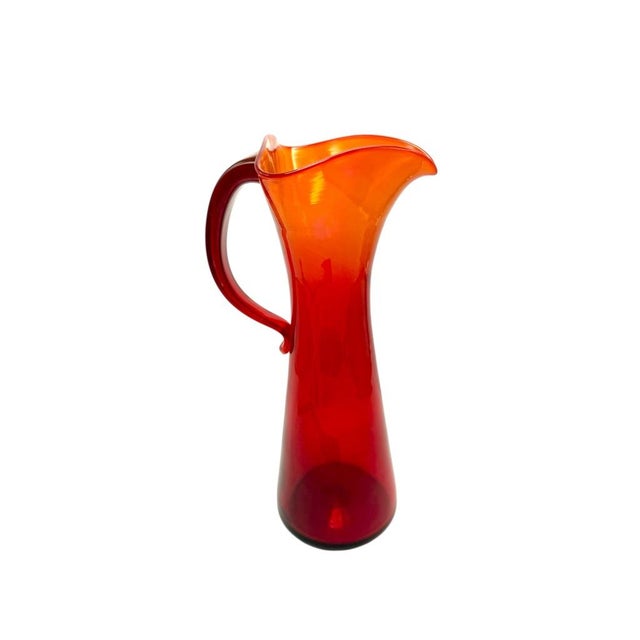 A striking mid-century modern hand-blown glass pitcher featuring a graceful silhouette with vibrant orange-to-red ombré...