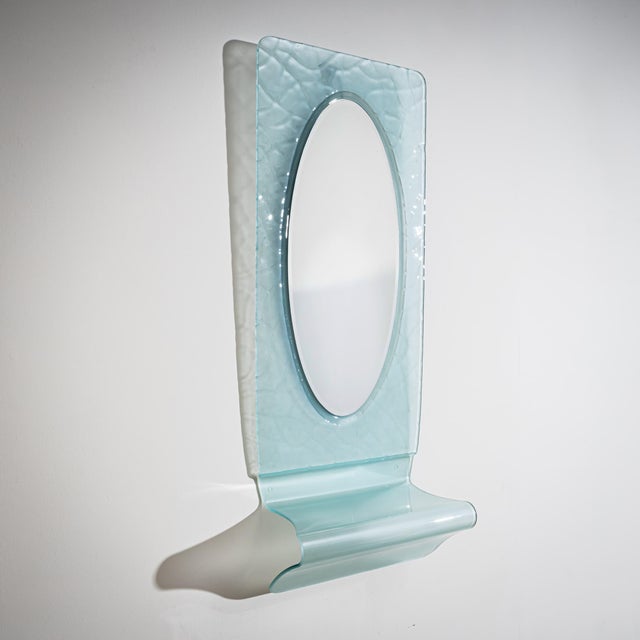 Oval wall mirror set within a rectangular frame with rounded corners, executed in light blue glass with a textured,...