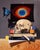 This oil on panel artwork presents a meticulously arranged still life featuring a human skull, children's blocks, and a...