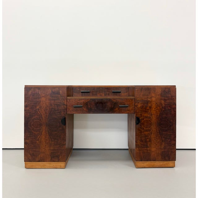 A streamlined Art Deco desk showcasing richly figured burl wood across its symmetrical pedestal form. Two central drawers...