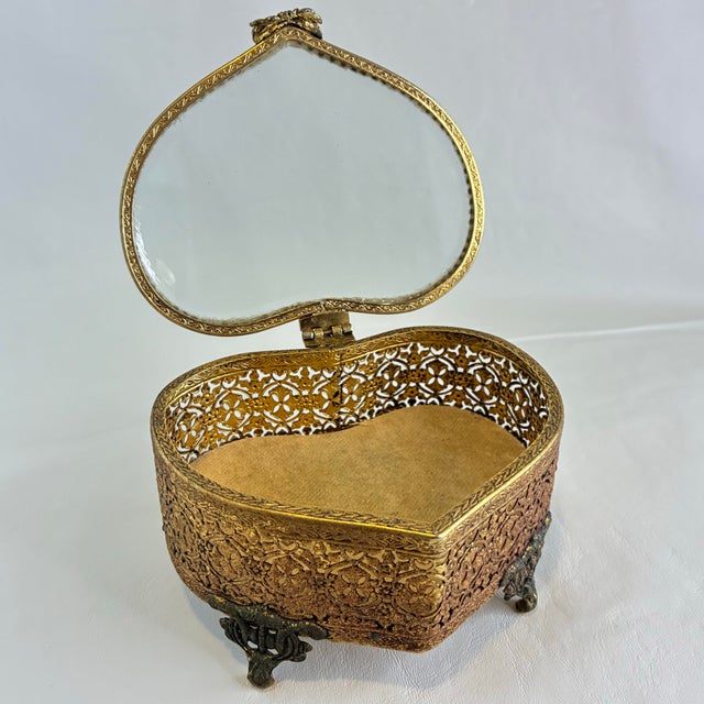 This vintage gold tone heart shaped jewelry box features an intricate ormolu filigree design with a clear glass lid....