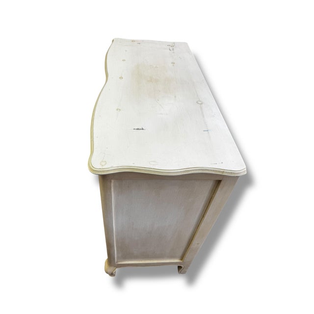A charming vintage dresser with graceful curves and timeless character, this piece features a softly distressed cream...