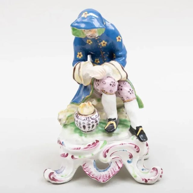 Antique 18th century Bow porcelain figure emblematic of Winter. The figure leaning before a fire and dressed in a bright...
