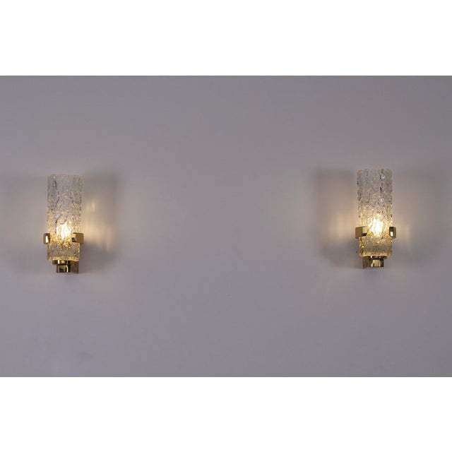 Pair of brutalist wall sconces, each consisting of a deeply engraved, gilded brass base supporting a thick, frosted glass...