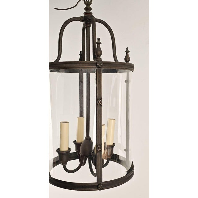 This elegant lantern features a cylindrical glass enclosure held within a bronze frame with gracefully arched supports and...