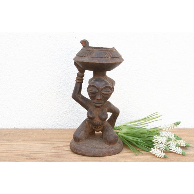 This elegant African figure, carved from wood with a rich, dark patina, depicts a woman kneeling with a vessel balanced...