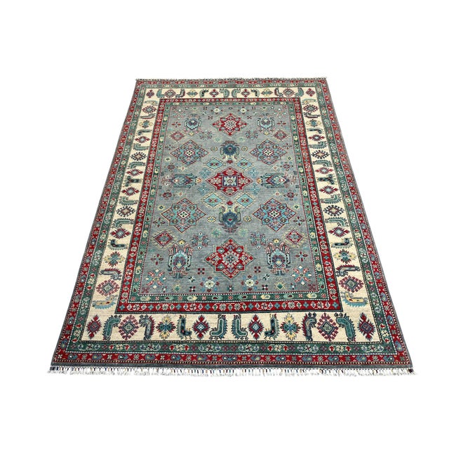 This Kazak-style wool area rug features a soft blue-gray field with bold red medallions and smaller geometric tribal...