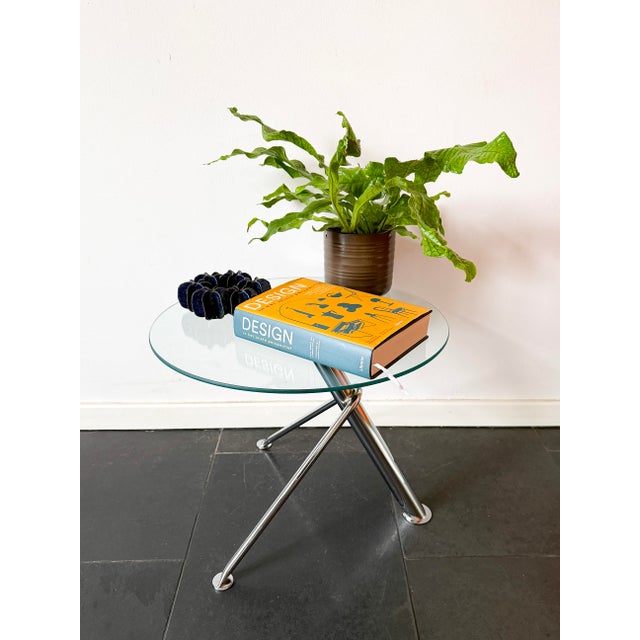 A round glass side table with a sculptural chrome-finished metal base, characterised by its minimalist modern design and...