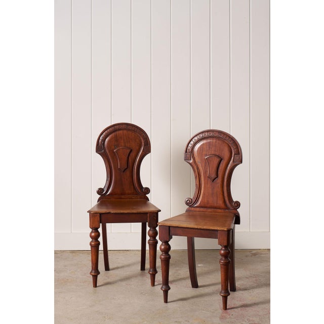 Pair of Victorian hall chairs with carved floral backs, shaped seats, and a warm aged finish. Classic English design with...