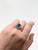 A blue zircon rests in a brilliant electric blue at the heart of this solid 14K rose gold ring, framed by forty-two earth-...