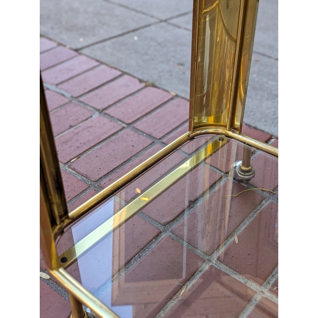 Vintage two-tier bar cart with a tubular brass frame, wide curved brass corner panels, and two clear glass shelves, on...