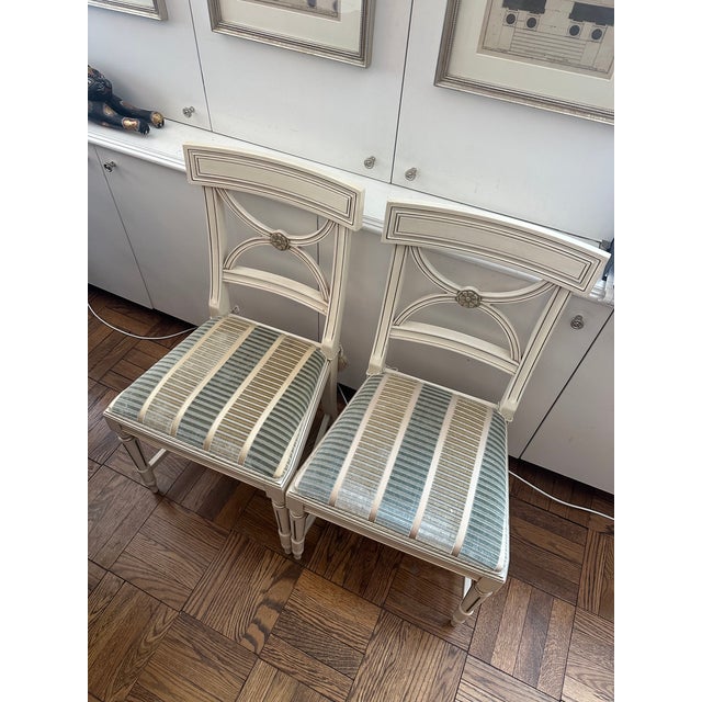 Two Bellman Chairs in white lacquered with gold gilding through-out chair. The seats are upholstered in S. Harris silk...