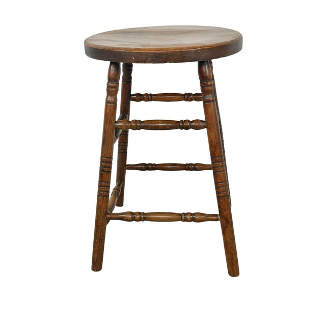 A charming antique country stool featuring a thick circular wooden seat, four elegantly turned legs, and an elaborate...