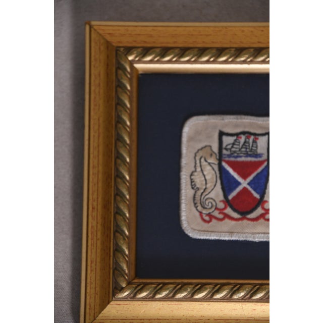 A beautifully framed embroidered patch featuring a ship crest flanked by seahorses on a navy linen mat, set in a gold...