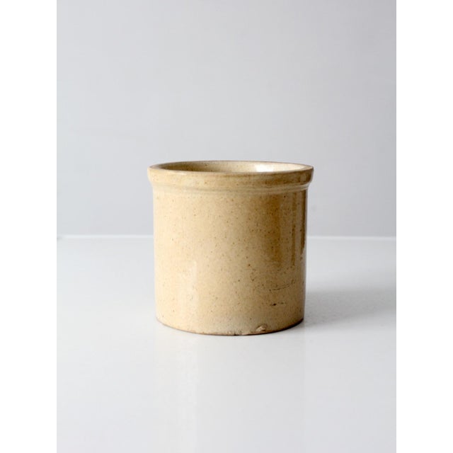 This antique stoneware crock features a simple cylindrical form with a substantial rolled rim and a warm cream-colored...