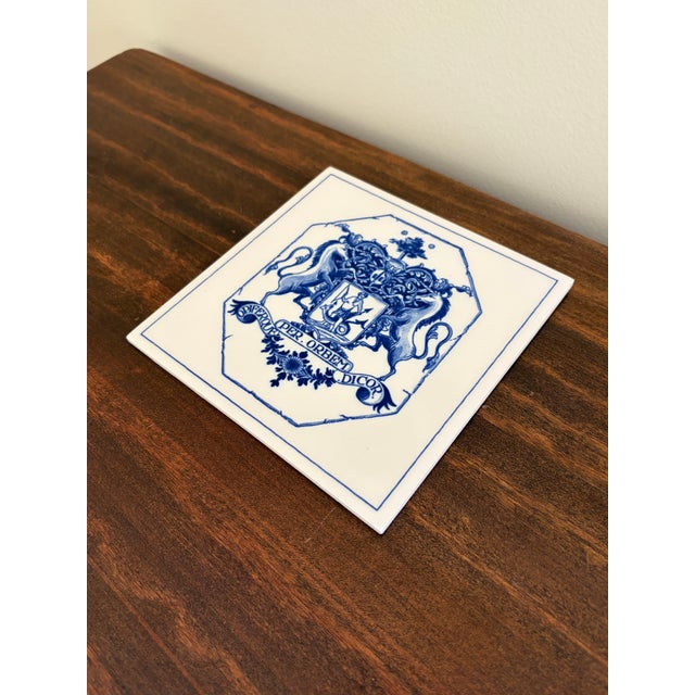 A classic Dutch Delft-style hand painted blue and white porcelain tile featuring an intricately rendered heraldic coat of...
