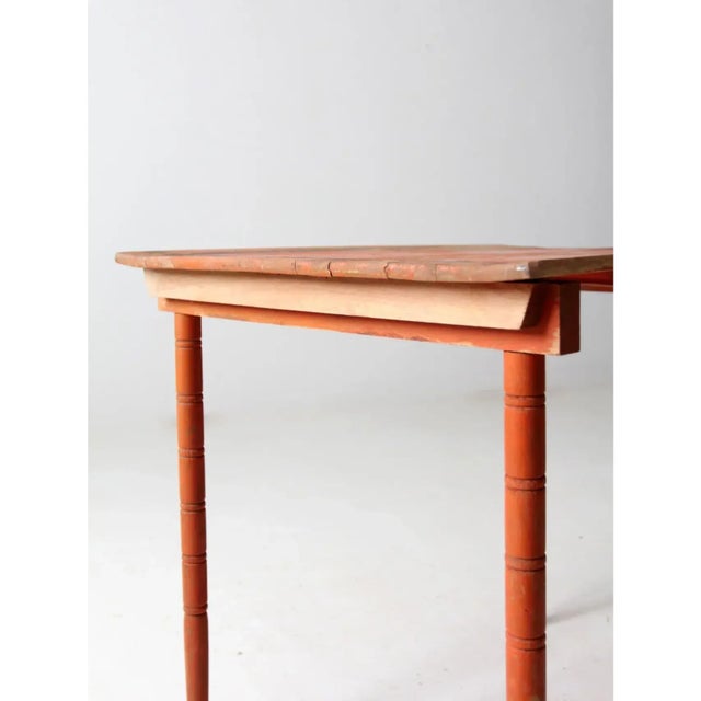 This antique “clipper” style folding table features a bold painted finish in warm orange with contrasting black accents...