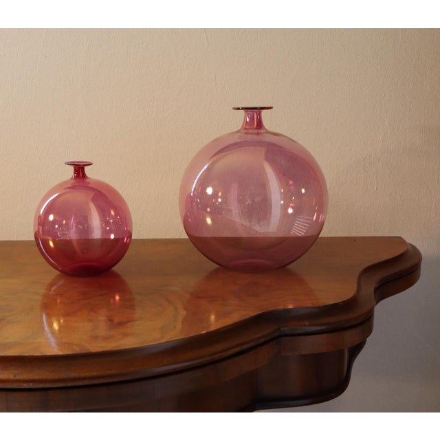 A pair of two amaranth pink blown glass vases in varying sizes. The vases are identical but different sizes. They are...