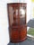 This elegant corner cabinet is crafted from wood, solid mahogany, mahogany veneer, and glass. It is labeled as being made...