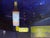 This oil on panel work captures a nighttime urban scene, where the glow of neon signs and a prominent bottle of Wild...