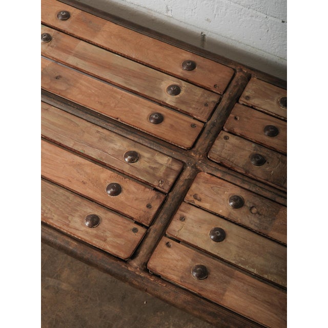 This iron and wood bench features a long, narrow profile with a slatted wooden seat secured by exposed metal rivets,...