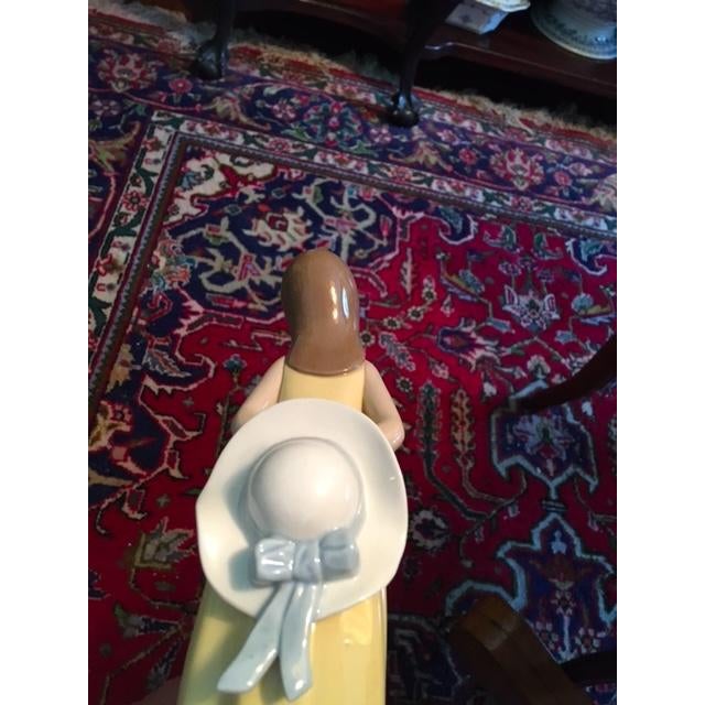 Pretty little girl figurine wearing a yellow dress with a red bow. Her white hat behind her has a blue ribbon. She has...