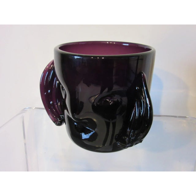 A purple hand blown glass head vase with a stylized face and hair in the manner of the Blenko glass company. This piece...