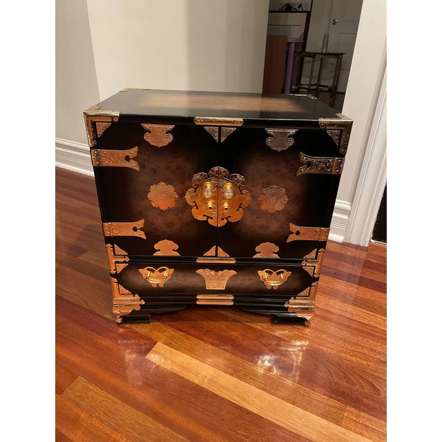 Striking 1980s burnished black lacquer cabinet featuring engraved brass corner mounts, decorative strap hinges, and ornate...