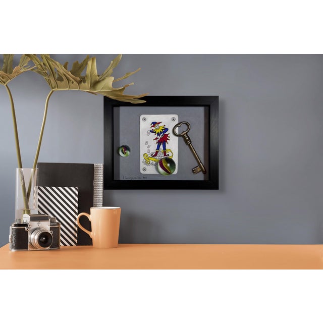 This artwork features a playful composition with a jester card, an antique key, and vibrant marbles, creating a sense of...
