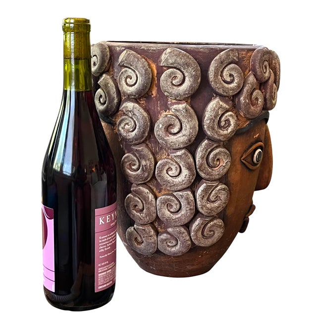 A striking handmade terra cotta vessel featuring a sculptural Janus-style double face motif, wrapped in dramatic applied...