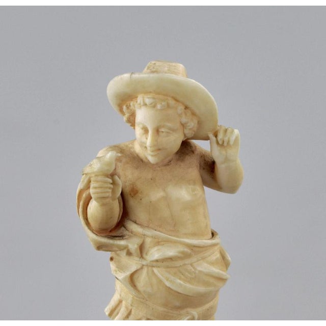 Carved ivory figurine of a boy with a bird, standing on a collapsible pedestal. A rural bird catcher in a straw hat is...