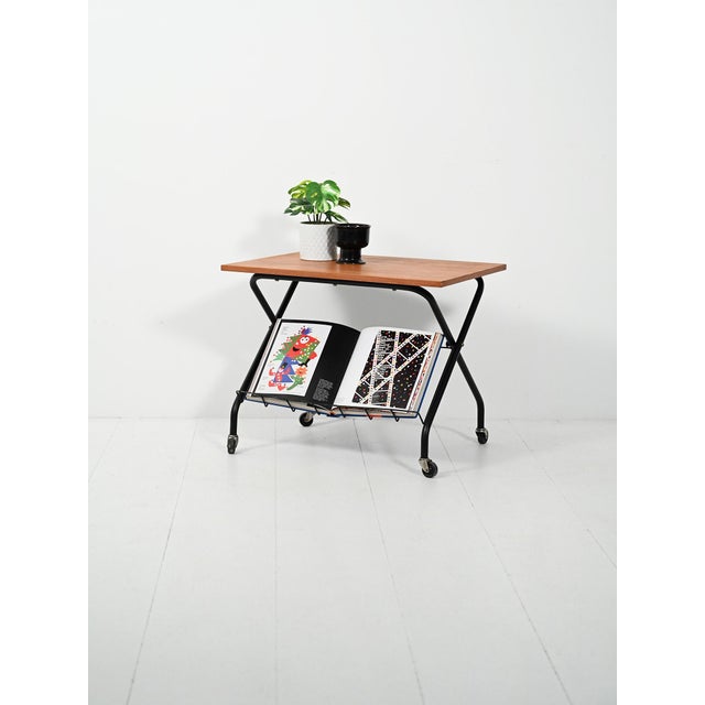 Teak and black-painted steel coffee table on casters, with a tubular X-shaped side frame and a metal wire magazine rack....