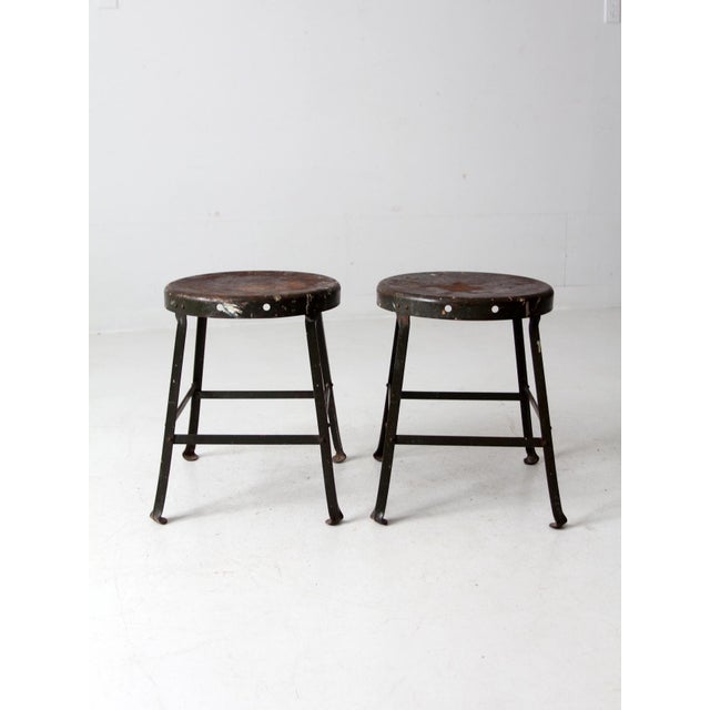 This is a pair of vintage industrial stools. The low shop stools feature dark green metal frames with round seats.