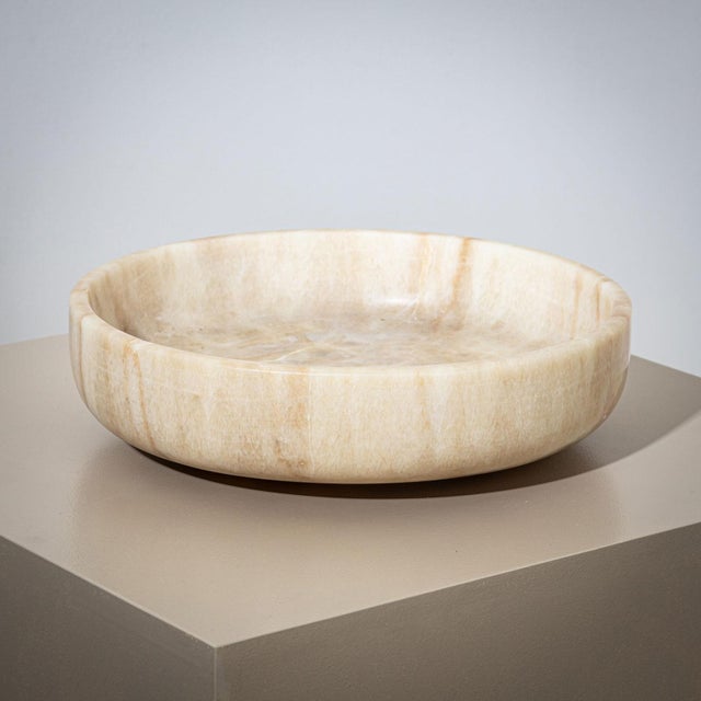 A large stone bowl crafted from beige onyx, featuring a minimalist design. The bowl is characterized by its high rim and...