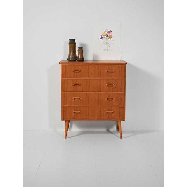 Teak chest of drawers with four drawers, double oval recessed handles, and obliquely turned legs. Soft lines, warm grain....
