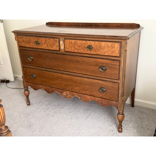 A wooden chest of drawers featuring two upper small drawers with burlwood veneer panels and a central carved rosette...
