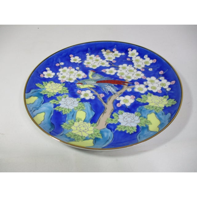 A decorative Asian porcelain plate with a gold trim. It is hand-painted with two blue and red pheasants resting on cherry...